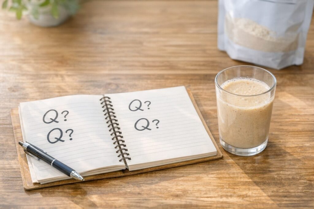ノートに書かれた複数の疑問符と、横に置かれたソイプロテインのグラスを俯瞰で撮影した横長写真