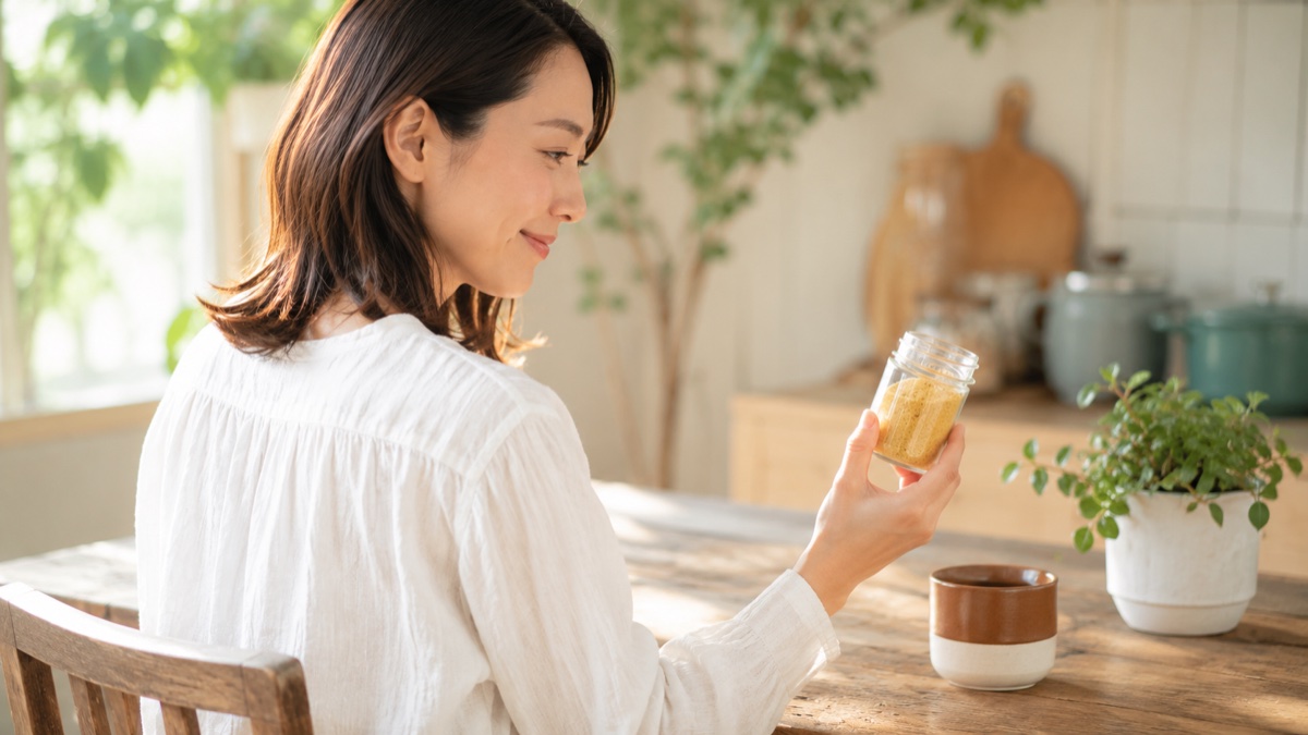 朝の自然光が差し込むナチュラルキッチンの木のテーブルで、コーヒーカップの横に置かれたアリサンのニュートリショナルイースト200gの粉末入り瓶を片手でそっと手に取って優しく眺める35歳の日本人女性。ヴィーガン9年の本音レビュー記事の導入シーン