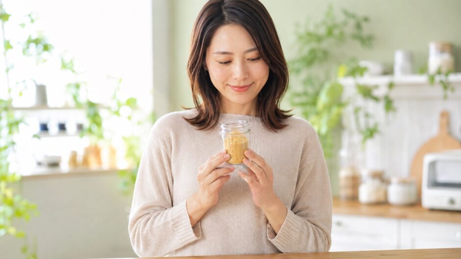 アリサンのニュートリショナルイースト200gの粉末入り瓶を両手で胸の前に持ち、ナチュラルキッチンを背景に優しく微笑む35歳の日本人女性。B12補給に役立つ栄養酵母の1年使用レビュー記事サムネイル