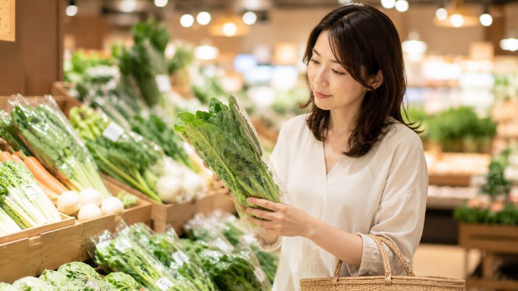 農薬を減らすために野菜を選んでいる場面。店頭の野菜売り場で、女性が葉物野菜を手に取り、どう育てられたかを確かめるように見ている