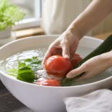 野菜の農薬を落とすために水を張ったボウル。朝の光が差すキッチンで、水に沈めたトマトやきゅうりを手がそっと押さえ、水面に光が揺れている