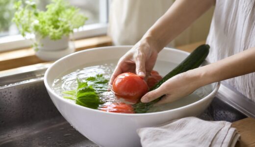 野菜の農薬を落とすために水を張ったボウル。朝の光が差すキッチンで、水に沈めたトマトやきゅうりを手がそっと押さえ、水面に光が揺れている
