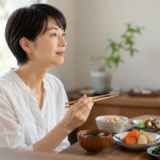 40代からの女性に足りていない栄養。明るいダイニングで、玄米や青菜、にんじん、わかめの汁ものなど品数の多い食事を前に、女性が箸を止めて考えている