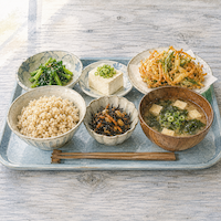 ヴィーガンダイエットの9年間、中身を変えていない食卓。玄米ごはんと味噌汁、青菜、にんじんとひじきの煮物、冷奴、揚げ物を青いトレーに並べた水平目線の水彩イラスト