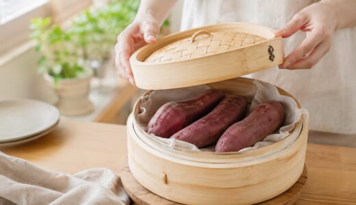 さつまいもを皮ごと蒸す場面。竹せいろの蓋を持ち上げた手元と、蒸し上がったばかりの皮つきのさつまいもが4本並ぶ。明るい自然光のキッチン
