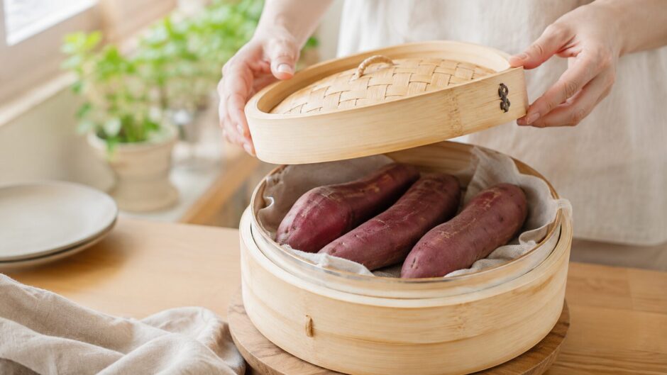 さつまいもを皮ごと蒸す場面。竹せいろの蓋を持ち上げた手元と、蒸し上がったばかりの皮つきのさつまいもが4本並ぶ。明るい自然光のキッチン