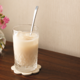 疲れた日に飲む甘酒の豆乳割り。氷を入れた背の高いカットグラスにスプーンを挿し、奥に百日紅のピンクの花を生けた小瓶を置いた我が家のテーブル