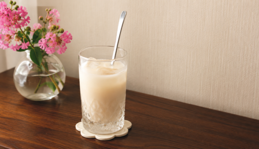 疲れた日に飲む甘酒の豆乳割り。氷を入れた背の高いカットグラスにスプーンを挿し、奥に百日紅のピンクの花を生けた小瓶を置いた我が家のテーブル