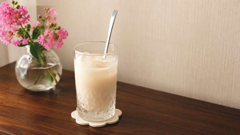 疲れた日に飲む甘酒の豆乳割り。氷を入れた背の高いカットグラスにスプーンを挿し、奥に百日紅のピンクの花を生けた小瓶を置いた我が家のテーブル