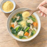 栄養酵母の粉末をスープにかける手元を描いた、ビタミンB12をとる食材を紹介する水彩イラスト