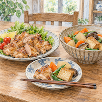ヴィーガンと家族の食事で勧めない食卓。焼いた鶏肉の大皿の隣に野菜と厚揚げの煮物の鉢が並び、手前の取り皿に両方が少しずつ取られている水彩イラスト