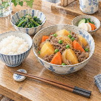 ヴィーガンと家族の食事と友人宅の料理。出されたばかりの湯気の上がる肉じゃがの鉢に、ほうれん草のごま和えと漬物、ごはんが並び、箸置きに箸がそろう水彩イラスト