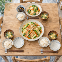 ヴィーガンと家族の食事を2種類に分けた食卓。真ん中の大皿の豚肉の野菜炒めを2人で分け、1人分だけ別の植物性の料理が並ぶ昼の水彩イラスト
