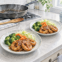 ヴィーガンと家族の食事で夫婦は同じ料理。台所の調理台に、車麩の照り焼きと野菜を盛った同じ皿が2枚並び、湯気が上がる水彩イラスト
