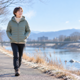 ヴィーガン視点で見るパタゴニアのダウン代替。冬の朝、ロゴのない軽い中綿ジャケットで川沿いを歩き、ほほえむ日本人女性