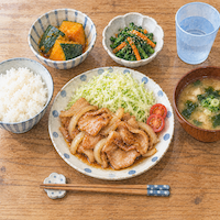 ヴィーガンと家族の食事で子どもの分に一品足す。真上から見た子どもの席に、夫婦と同じ味噌汁やかぼちゃの煮物、サラダと、生姜焼きの皿が1つだけ多く並ぶ水彩イラスト