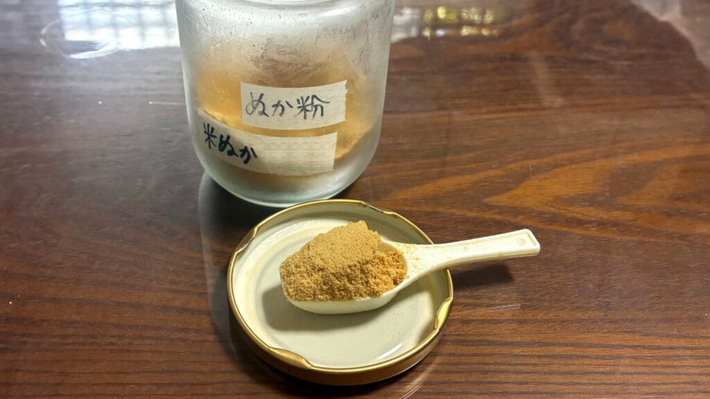 毎日使っている米ぬかパウダーの手書きラベルのガラス瓶と、瓶のふたの上で山盛りにした小さなスプーン1杯(約5g)